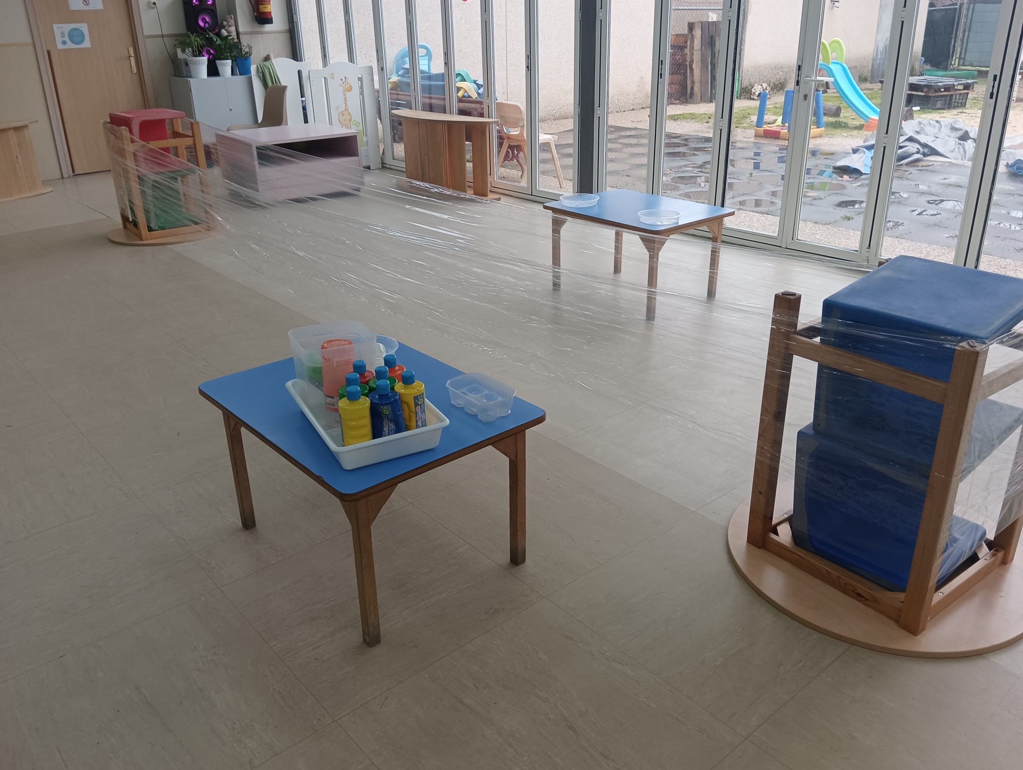 Estructura de madera con papel film transparente y botes de pintura preparada para una actividad sensorial infantil.