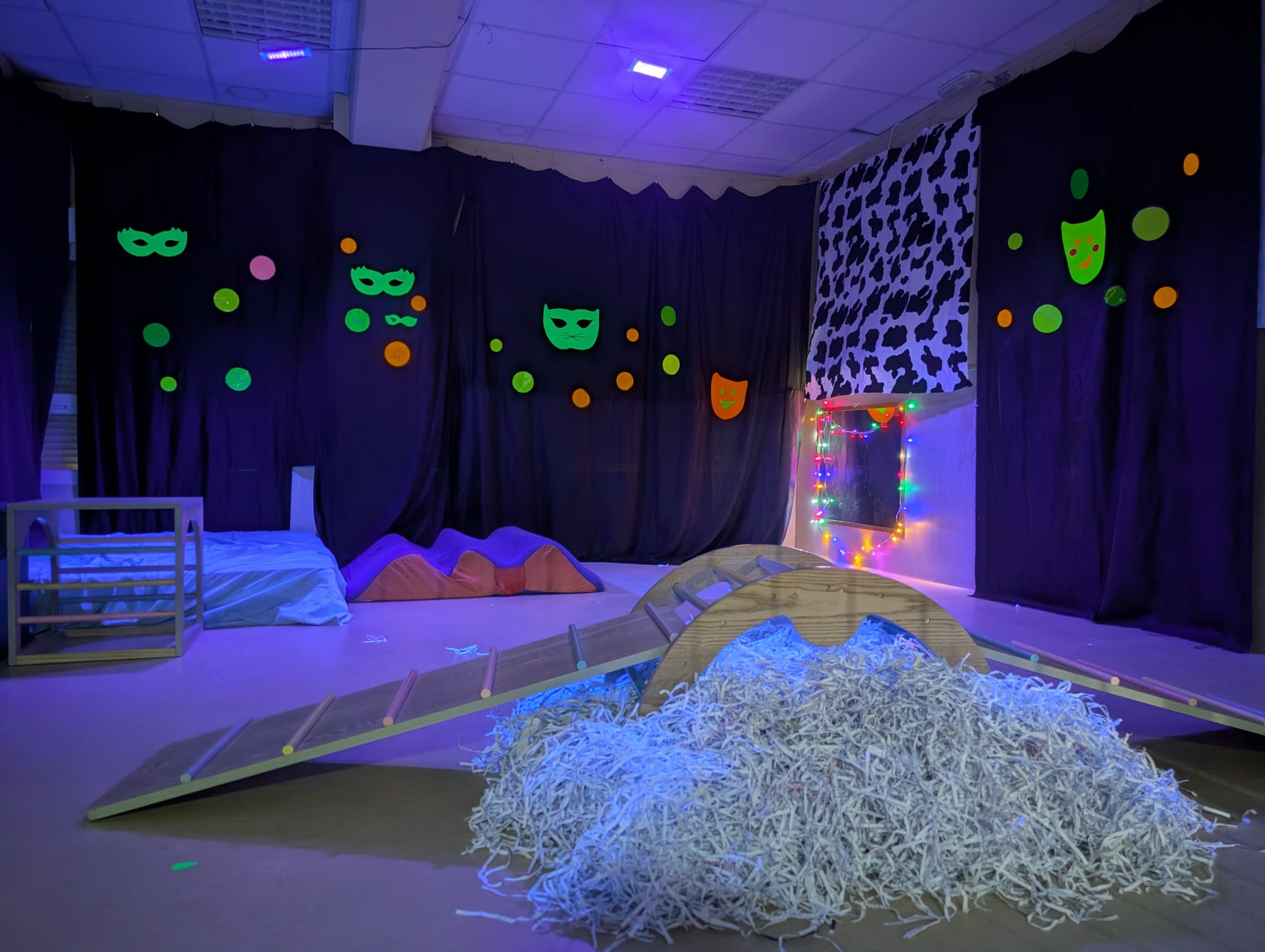 Vista general de una sala oscura decorada con máscaras de neón, luces de colores, rampas de madera y elementos de psicomotricidad.